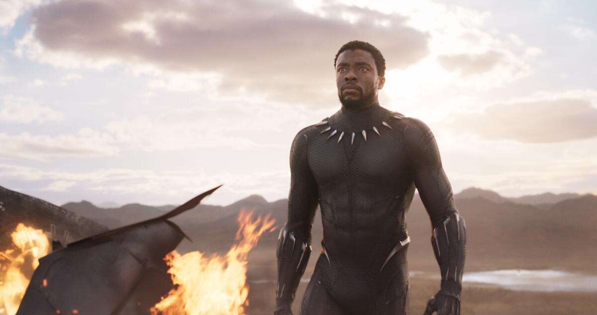 A man in a black superhero suit walks past flaming wreckage.