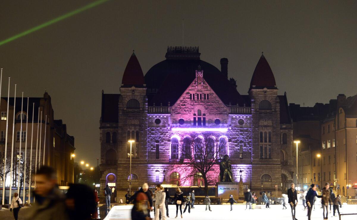 A pink and purple light installation on an imposing building in Helsinki, Finland, with people skating in front of it.