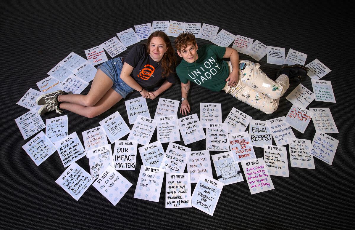 Two women lie on their sides on the floor, surrounded by dozens of papers with handwritten messages on them