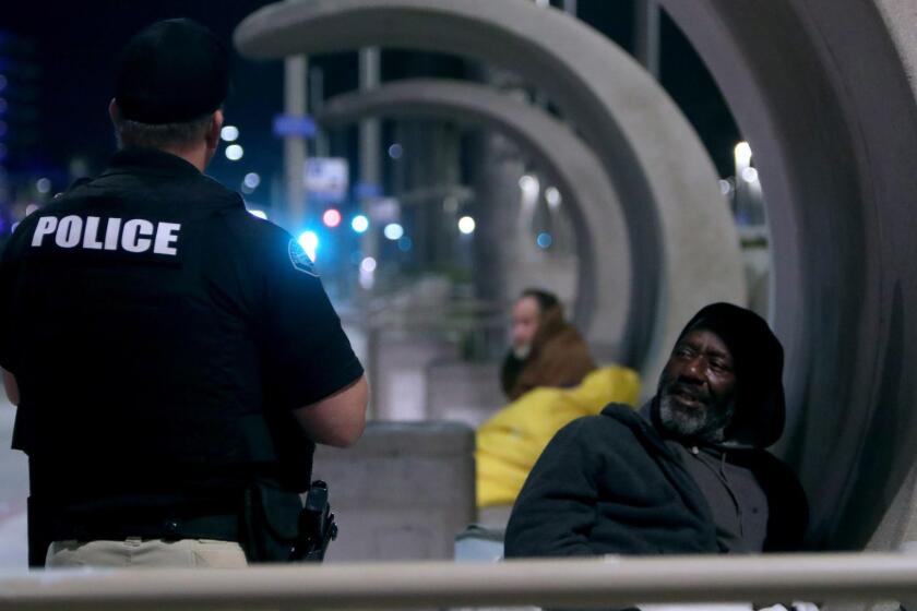 Huntington Beach Police Homeless Task Force officer Gabe Ricci speaks with a homeless man sleeping on a bench on Pacific Coast Highway in Huntington Beach, in the early hours of Wednesday, Feb. 13, 2019.