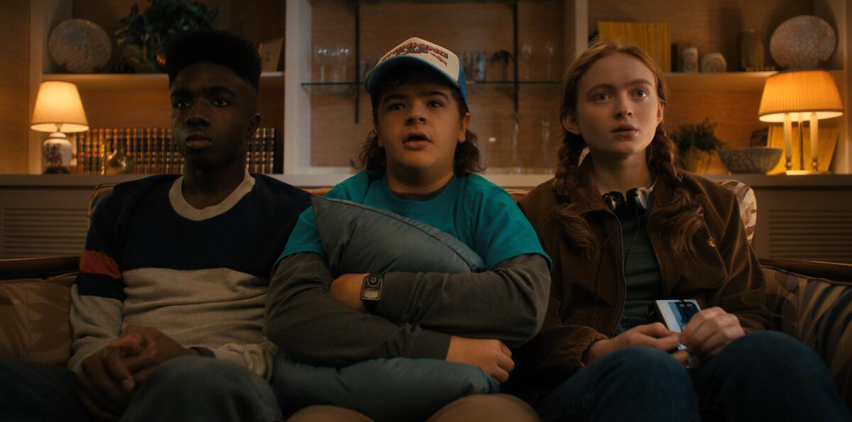 Three teens sit on a couch in a living room in a scene from Netflix's "Stranger Things."