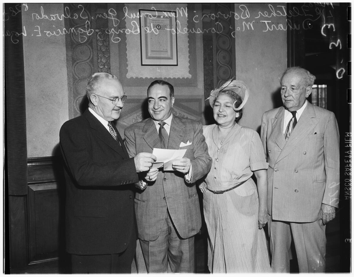 A black-and-white photo of three men and a woman standing and looking at a piece of paper held by two of the men