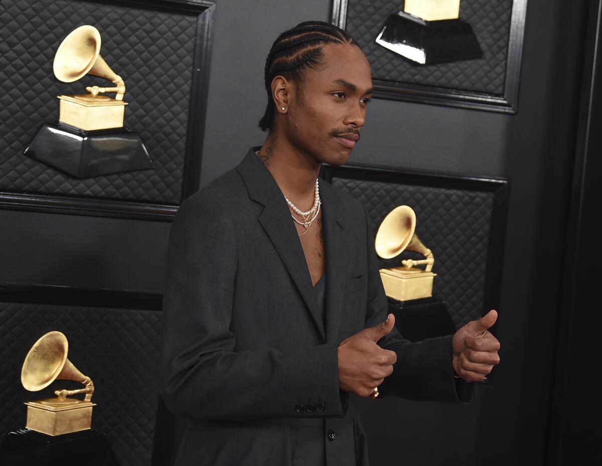 A man with braided black hair wearing a black suit giving two thumbs up in front of a Grammy backdrop