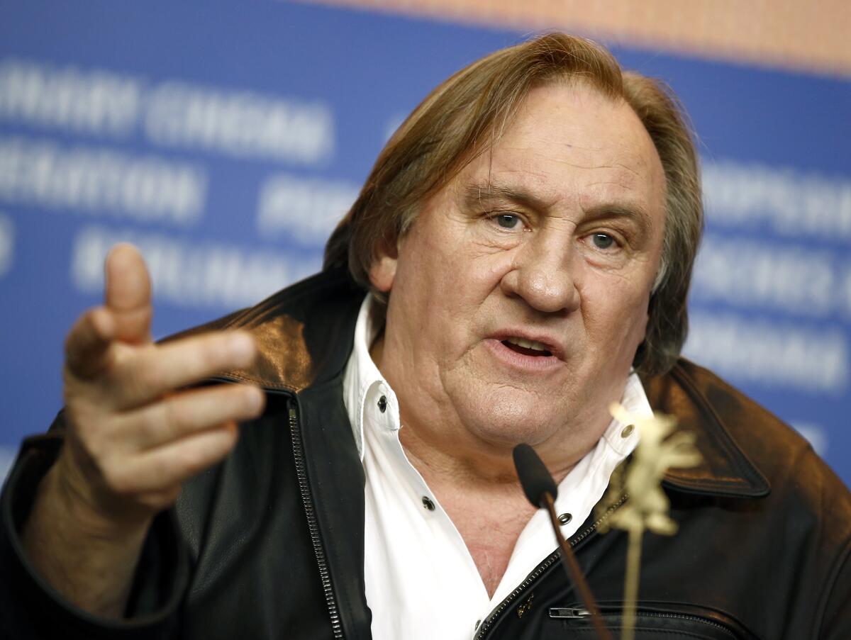 Actor Gerard Depardieu gestures with his hand while speaking in a brown jacket and white shirt behind a microphone