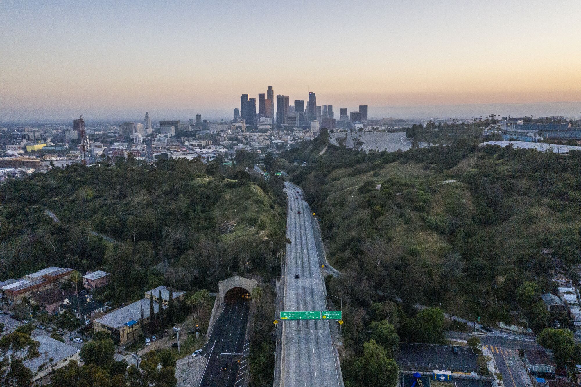 Aerial view of a mostly empty freeway running between green hills toward the downtown L.A. skyline