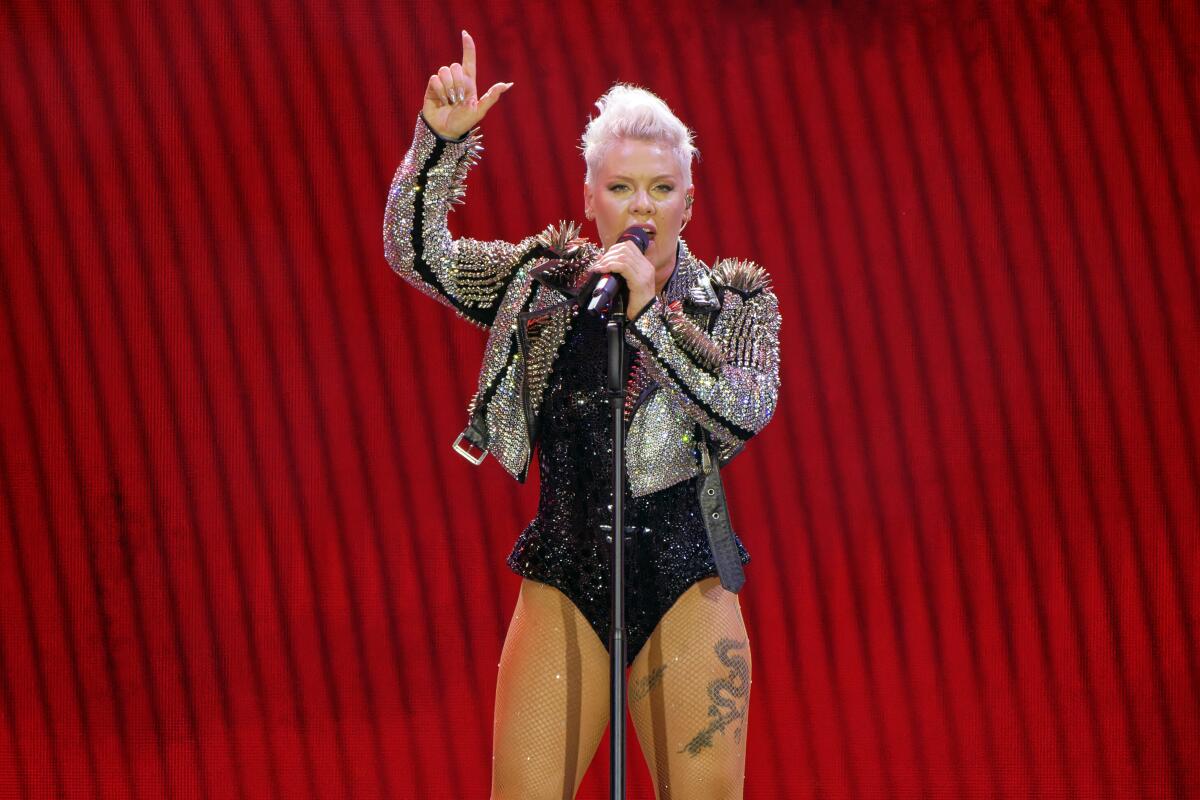 Pink in bedazzled in a leather jacket and leotard on stage speaking into a microphone and pointing upward with her right hand