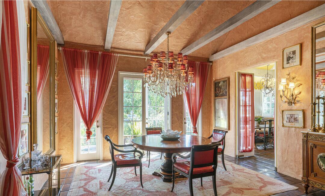 The dining area is in shades of orange with a dining table and chairs, a rug, art and windows overlooking foliage.