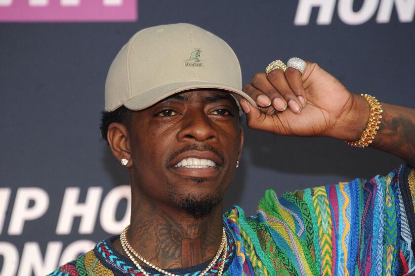 Rich Homie Quan holding the brim of a khaki hat. He smiles and wears a colorful sweater and chains