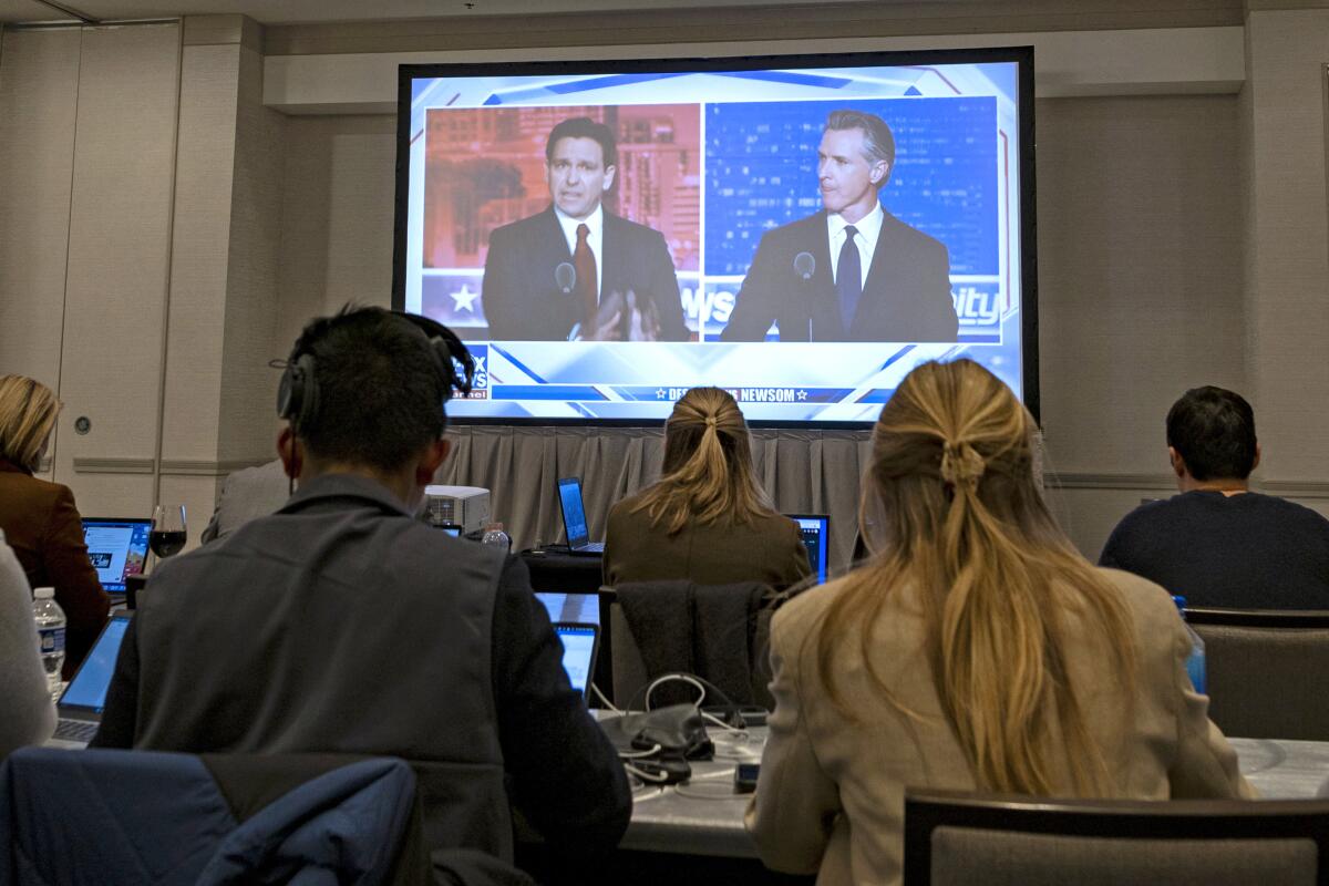 Side by side images of Florida Gov. Ron DeSantis and Gov. Gavin Newsom on a big screen with seated people facing them.