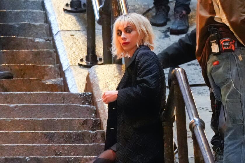 Lady Gaga dressed as Harley Quinn with white makeup and a dark coat leans against a railing along set of concrete stairs