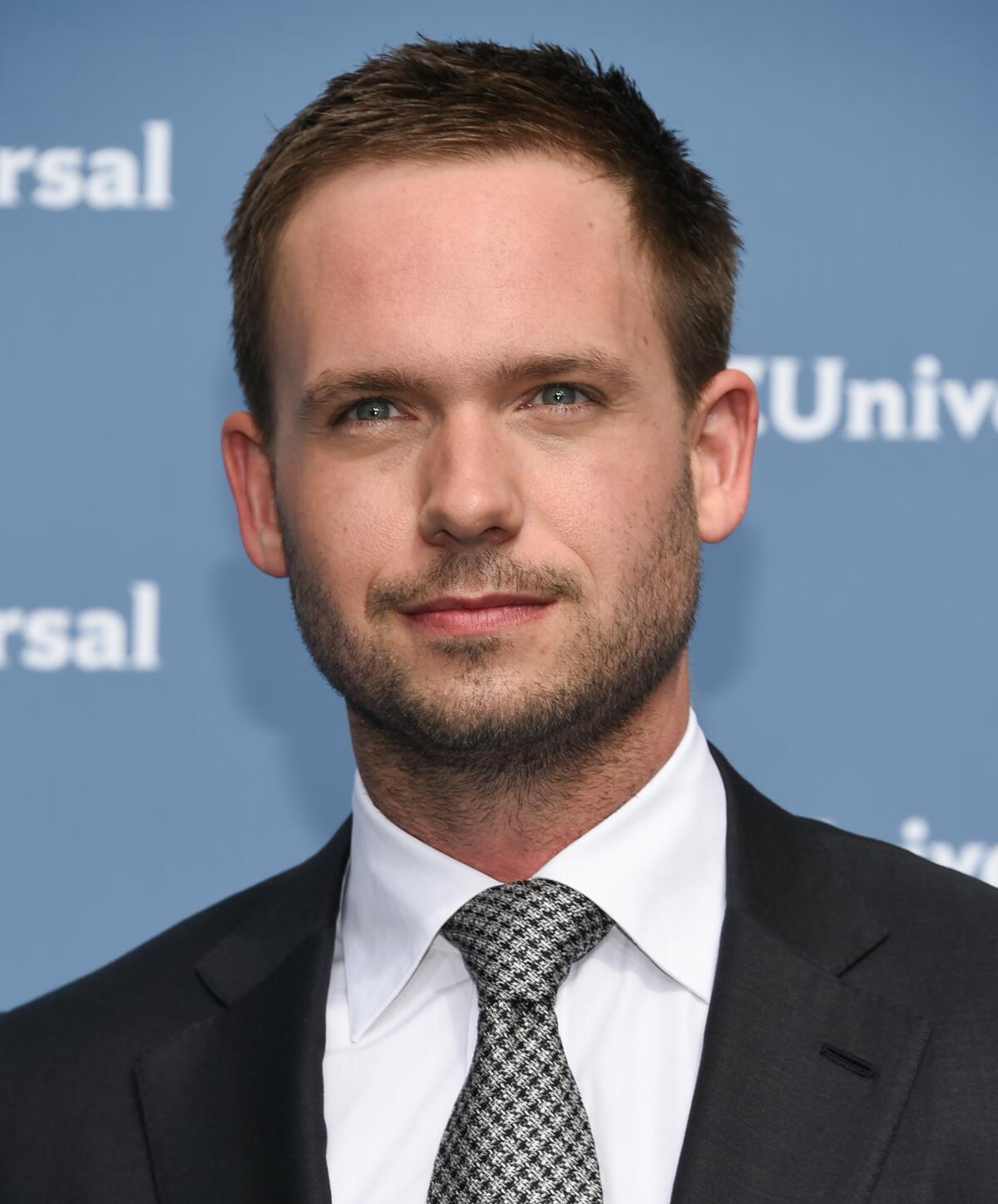 Actor Patrick J. Adams looks straight ahead in a dark suit, white shirt, silver patterned tie and facial stubble