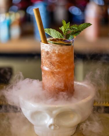A cocktail in a tall glass sits in a skull dish that oozes dry ice steam