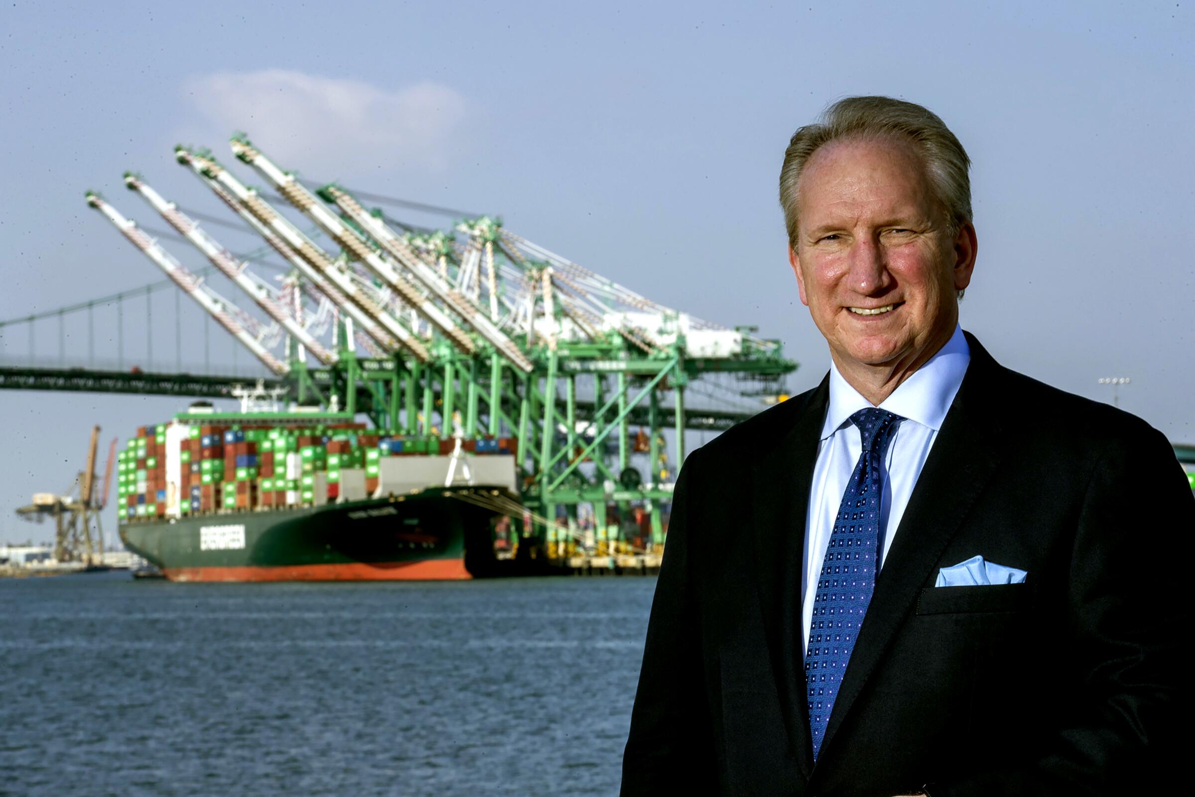 Gene Seroka standing in a black suit near the water with a container ship, cranes and a bridge in the distance
