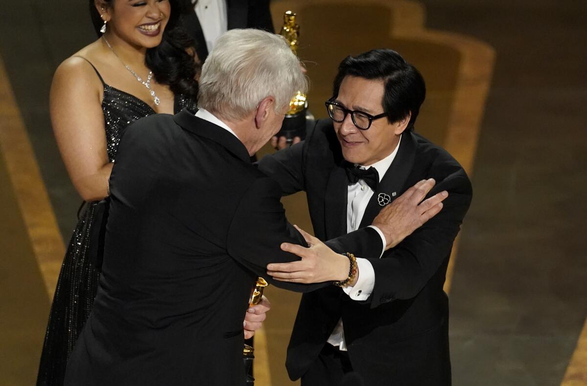 A man with white hair in a tuxedo embraces a man with black hair in a tuxedo on a stage