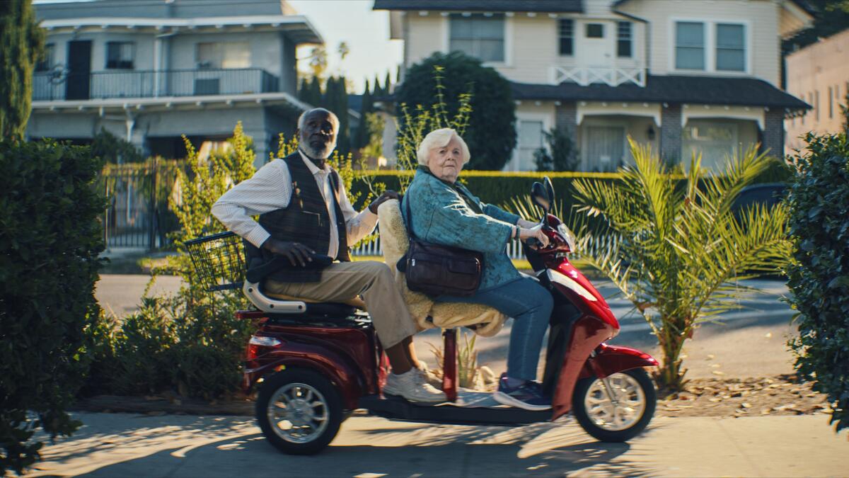 A gray-haired white woman drives a scooter with a Black man behind her as passenger