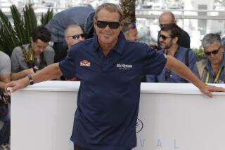 Chad McQueen in a blue shirt and shades posing for photographers with his arms spread wide on a tabletop behind him