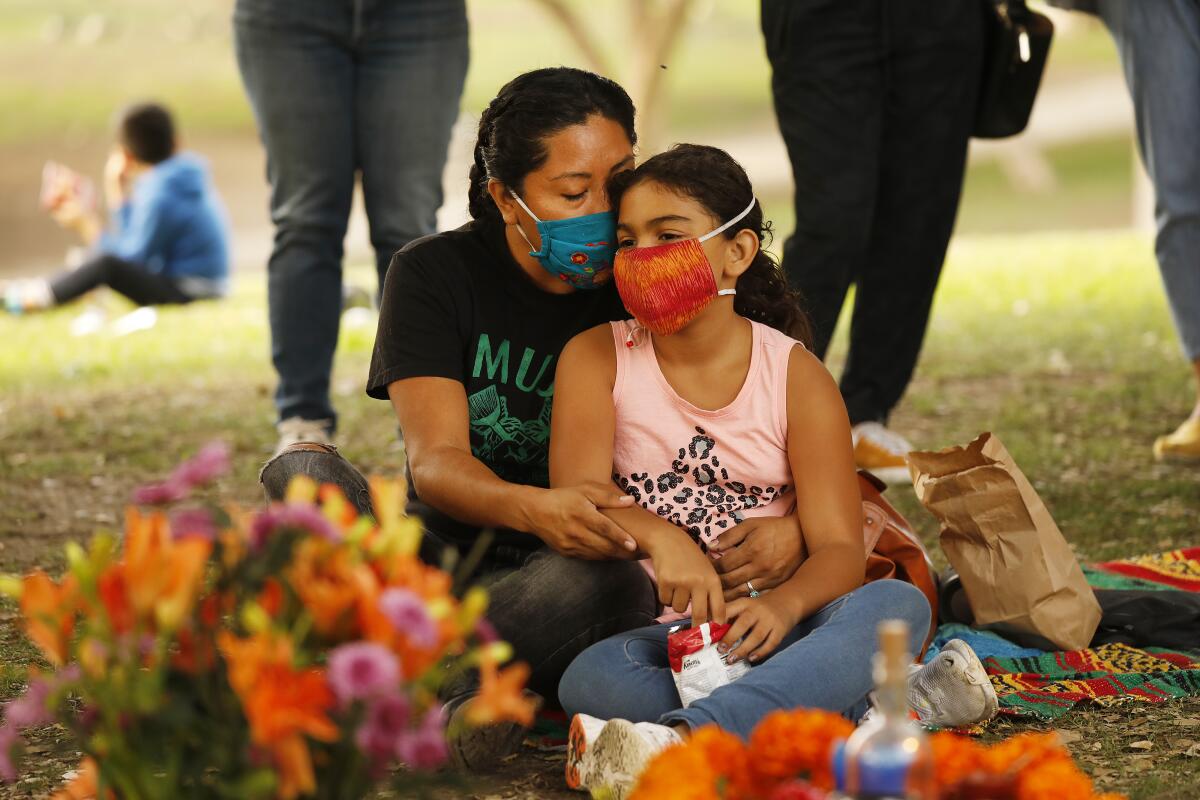 A woman holds her daughter as they both sit on the ground while wearing masks