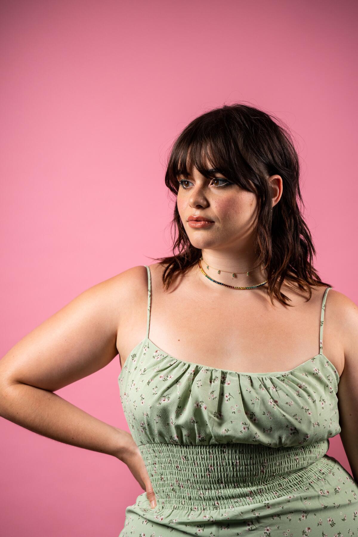 Barbie Ferreira poses, hand on one hip, in a green flowered, strappy sundress against a pink background.