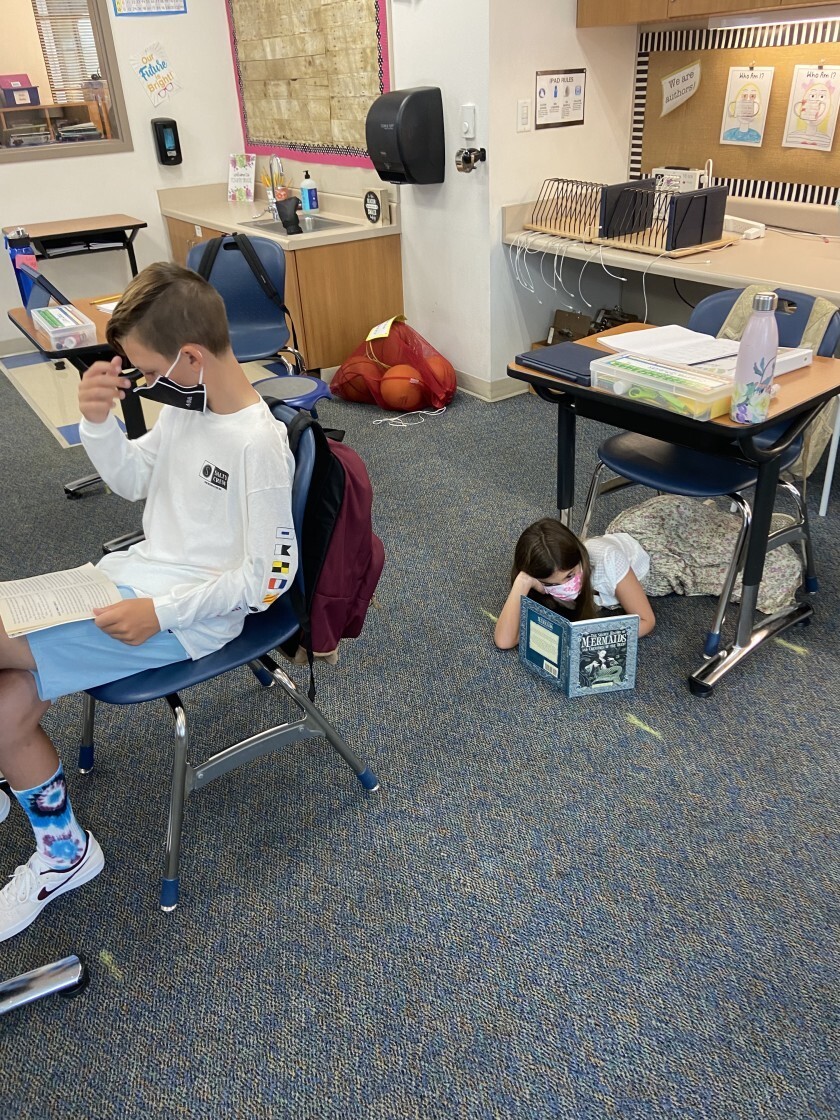 A masked boy, left, reads at his desk while a masked girl behind him reads lying on the floor under her desk