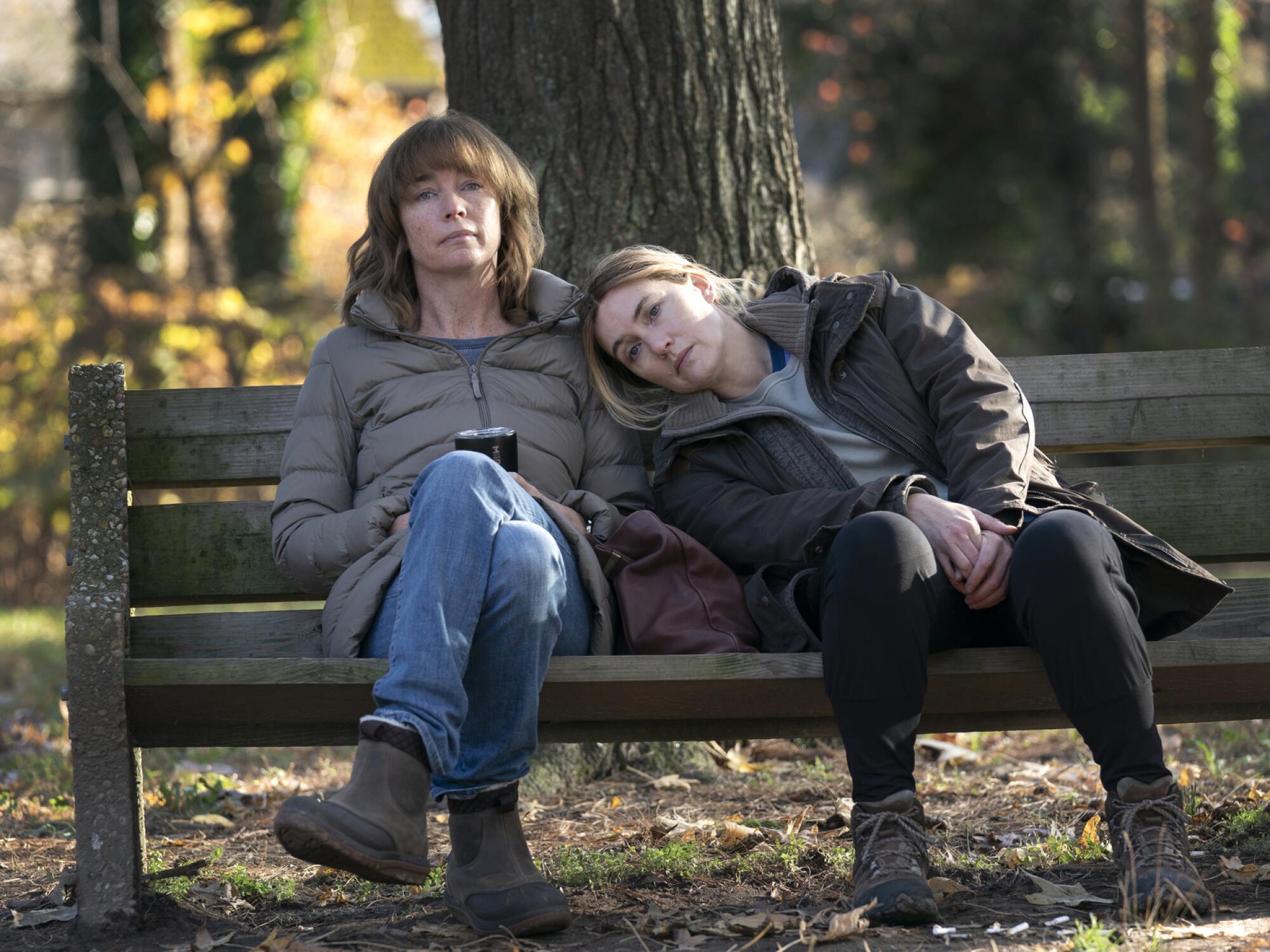 Two women sit on a park bench with one resting her head on the other's shoulder