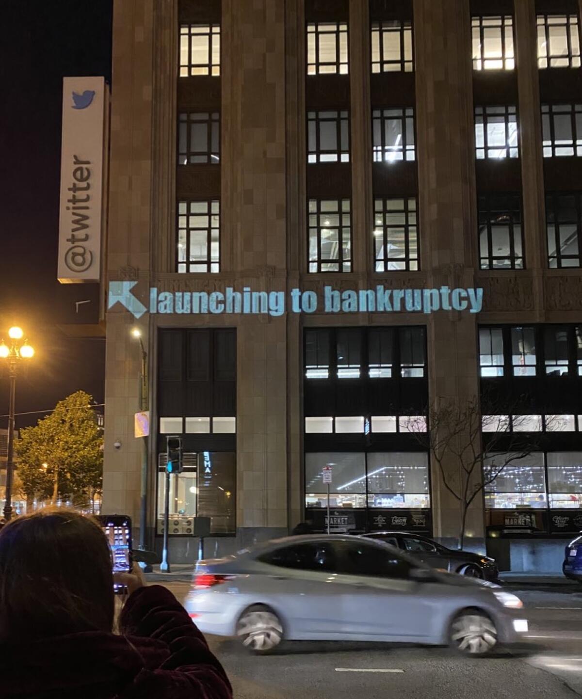 Large office building at night with "Launching to bankruptcy" projected on the front, pointing to a Twitter logo