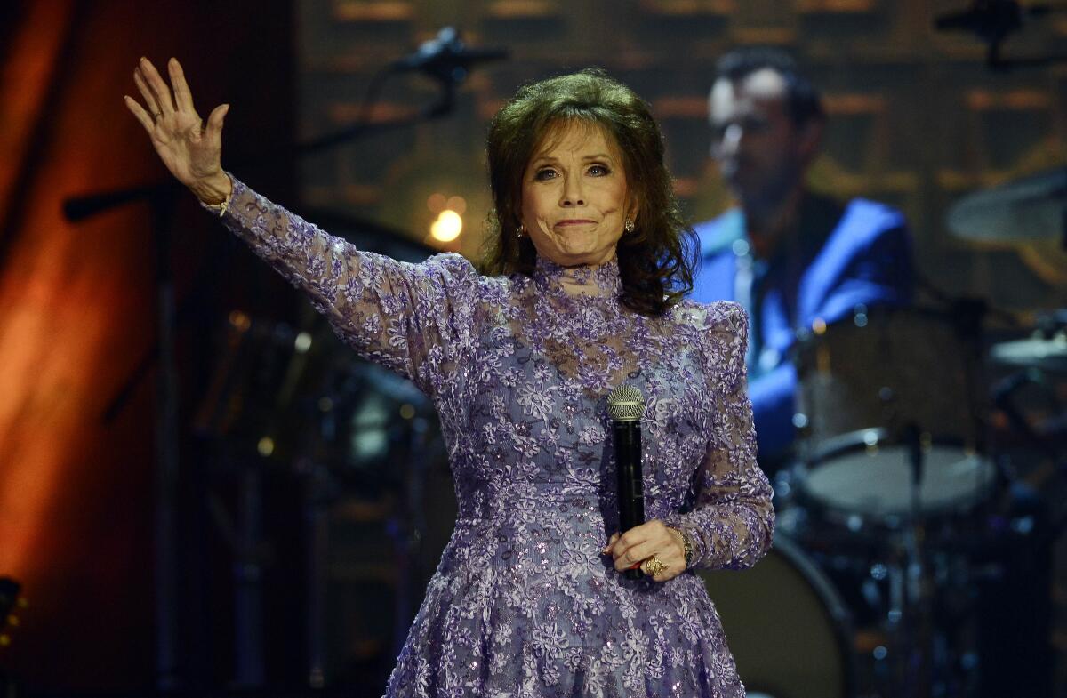 A woman with dark hair holding a microphone and waving from a stage in a long-sleeve floral lace gown