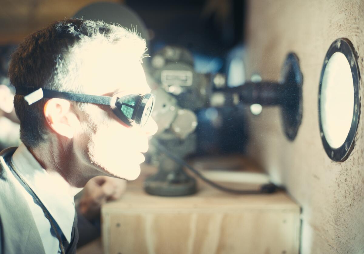 A man in dark goggles looks through a round window as a brilliant flash of light illuminates his face in "Oppenheimer."