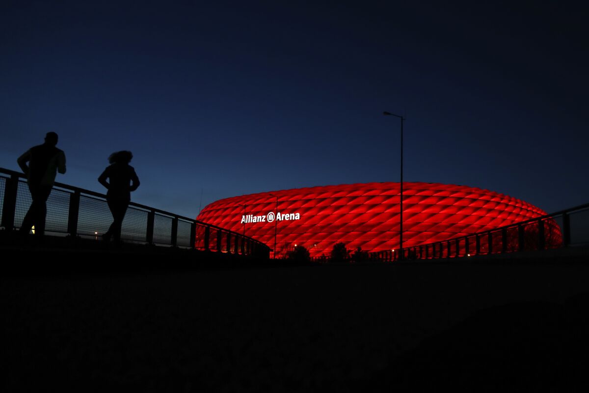 FILE-In this March 16, 2020 taken photo jogger make their way after the sun sets at the 'Allianz Arena' soccer stadium in Munich, Germany. European ruling body UEFA said on Friday the German city of Munich will remain as one of the Euro 2020 host cities. The confirmation came during a UEFA executive board videoconference after the federation was informed by local authorities that all four matches in Munich will be able to welcome a minimum of 14,500 spectators. (AP Photo/Matthias Schrader)