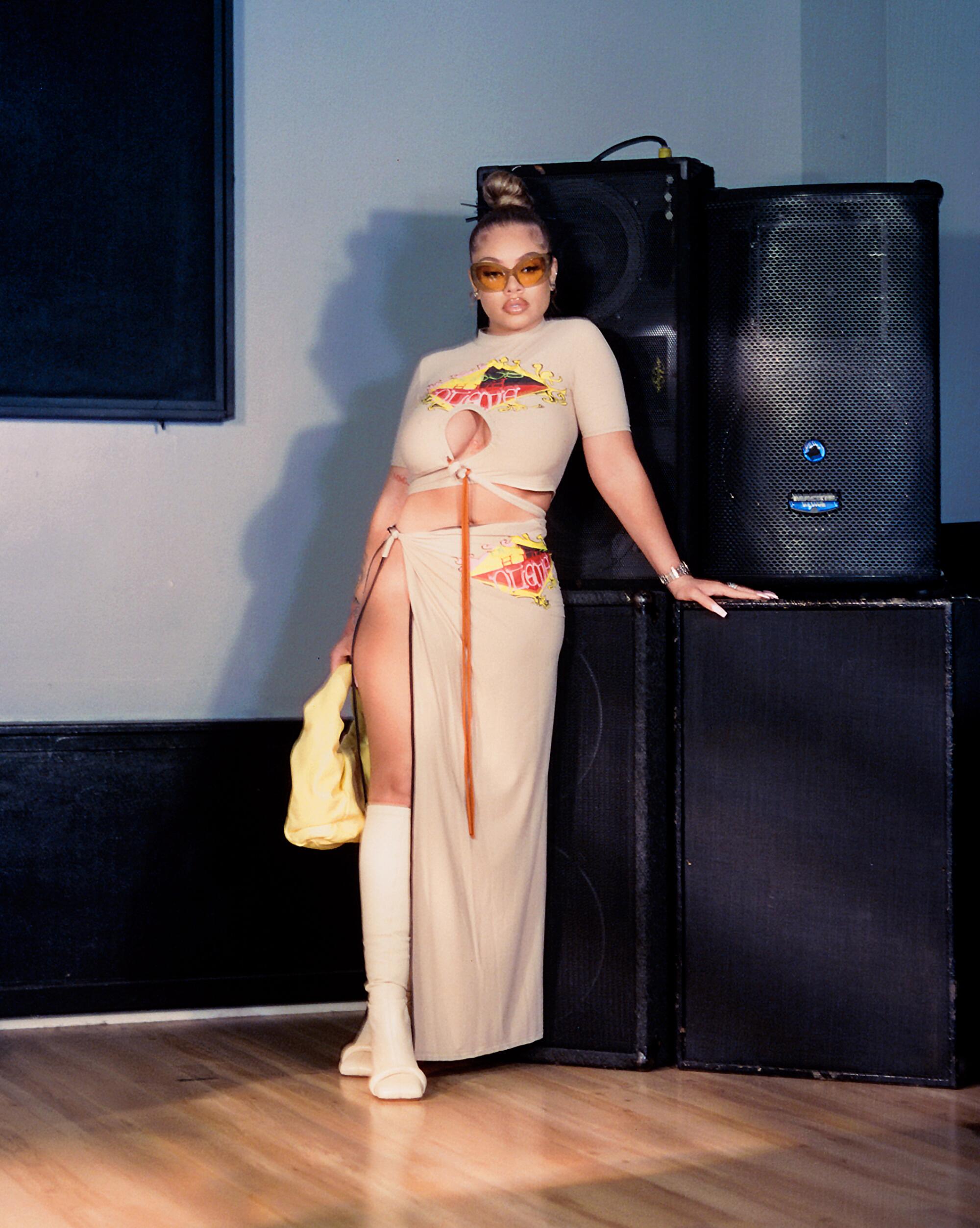A female rapper in a tan dress poses in front of large speakers