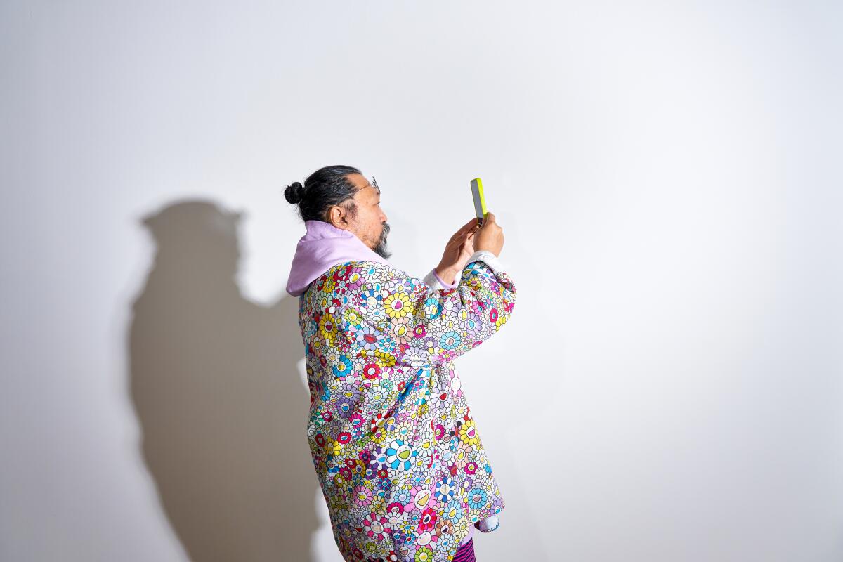 Takashi Murakami, in a hoodie covered in his cartoon flowers, is seen against a white wall looking at his phone.