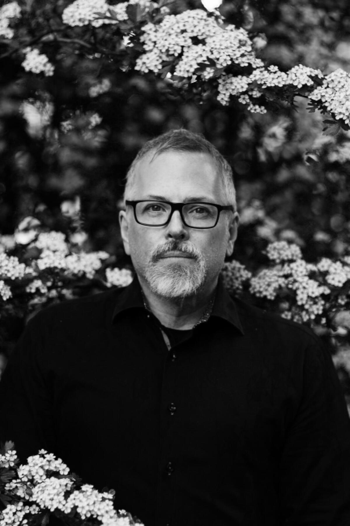 Jeff Vandermeer, with salt and pepper hair and trim beard in glasses and a dark shirt with flower blossoms behind him.