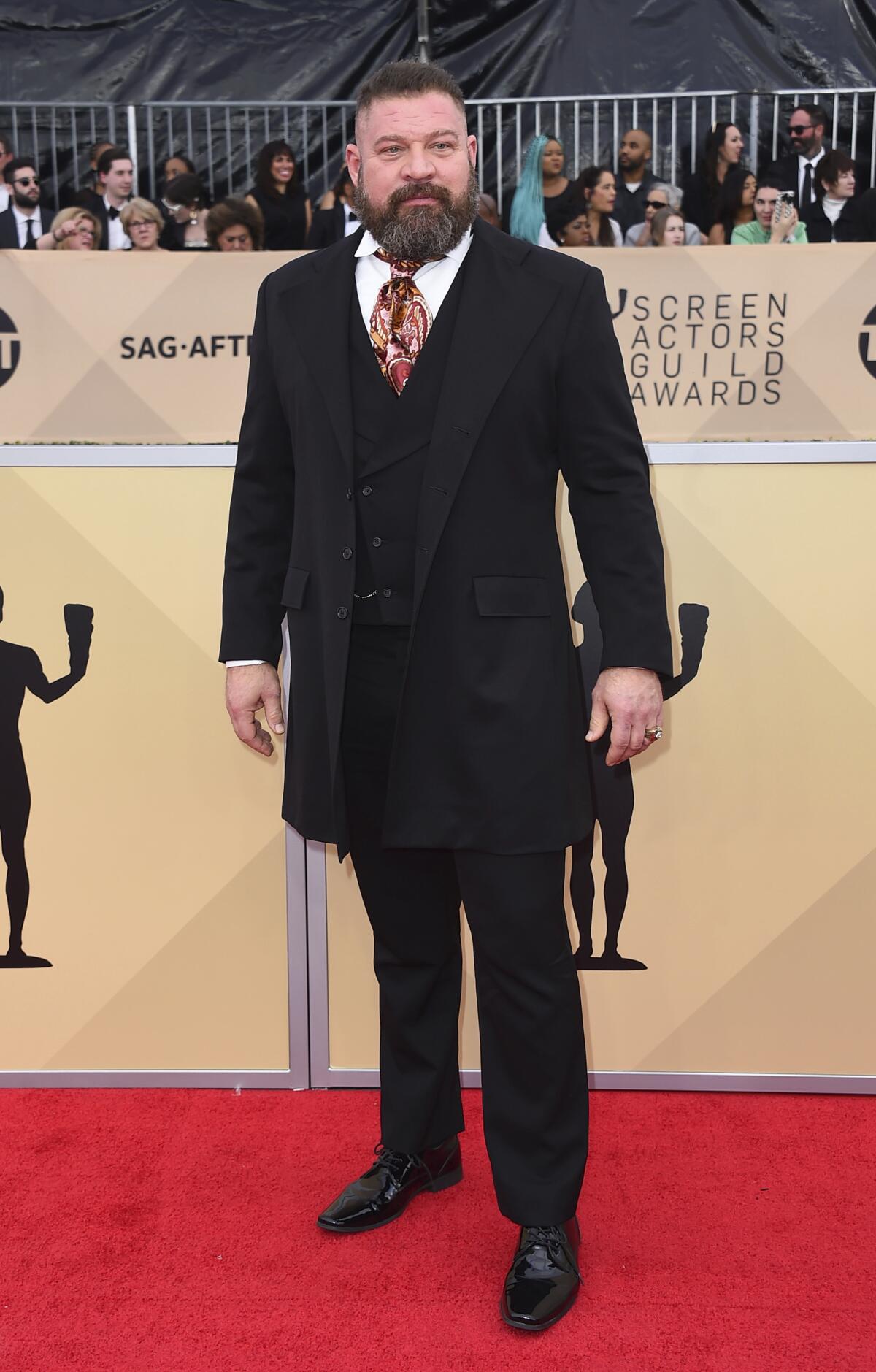 A tall man in a black three-piece suit stands on a red carpet