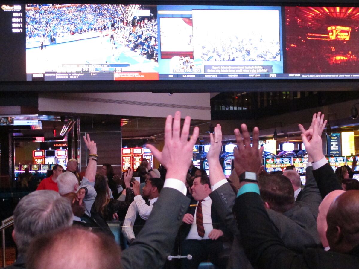 TV screens are seen overhead as a crowd of people raise up their hands.