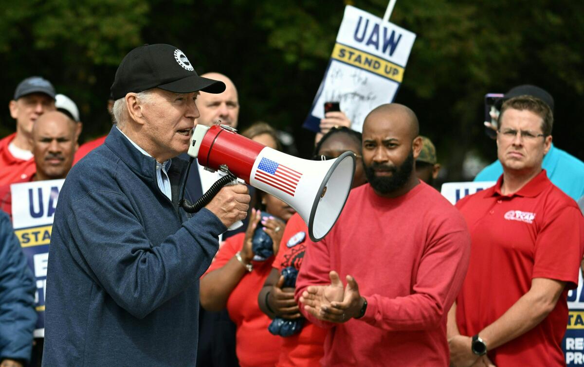 A man in a dark cap and jacket speaks into a bullhorn bearing a U.S. flag as some people in red shirts applaud 