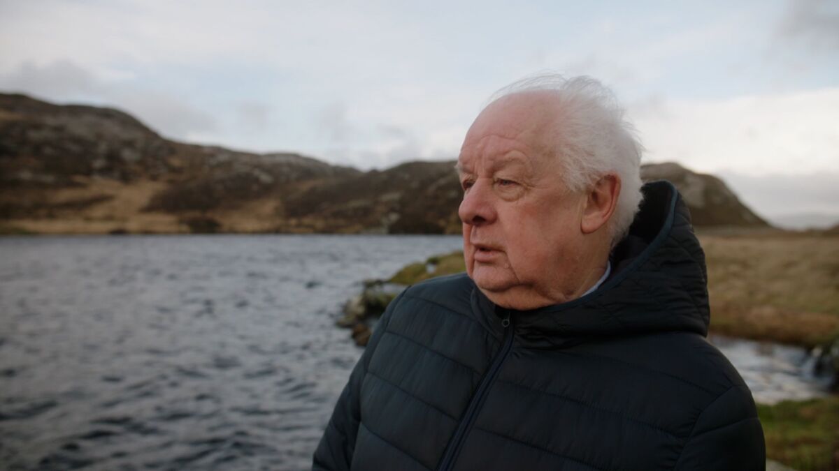 A man with gray hair and wearing a hooded jacket stands on a windy lake shore.