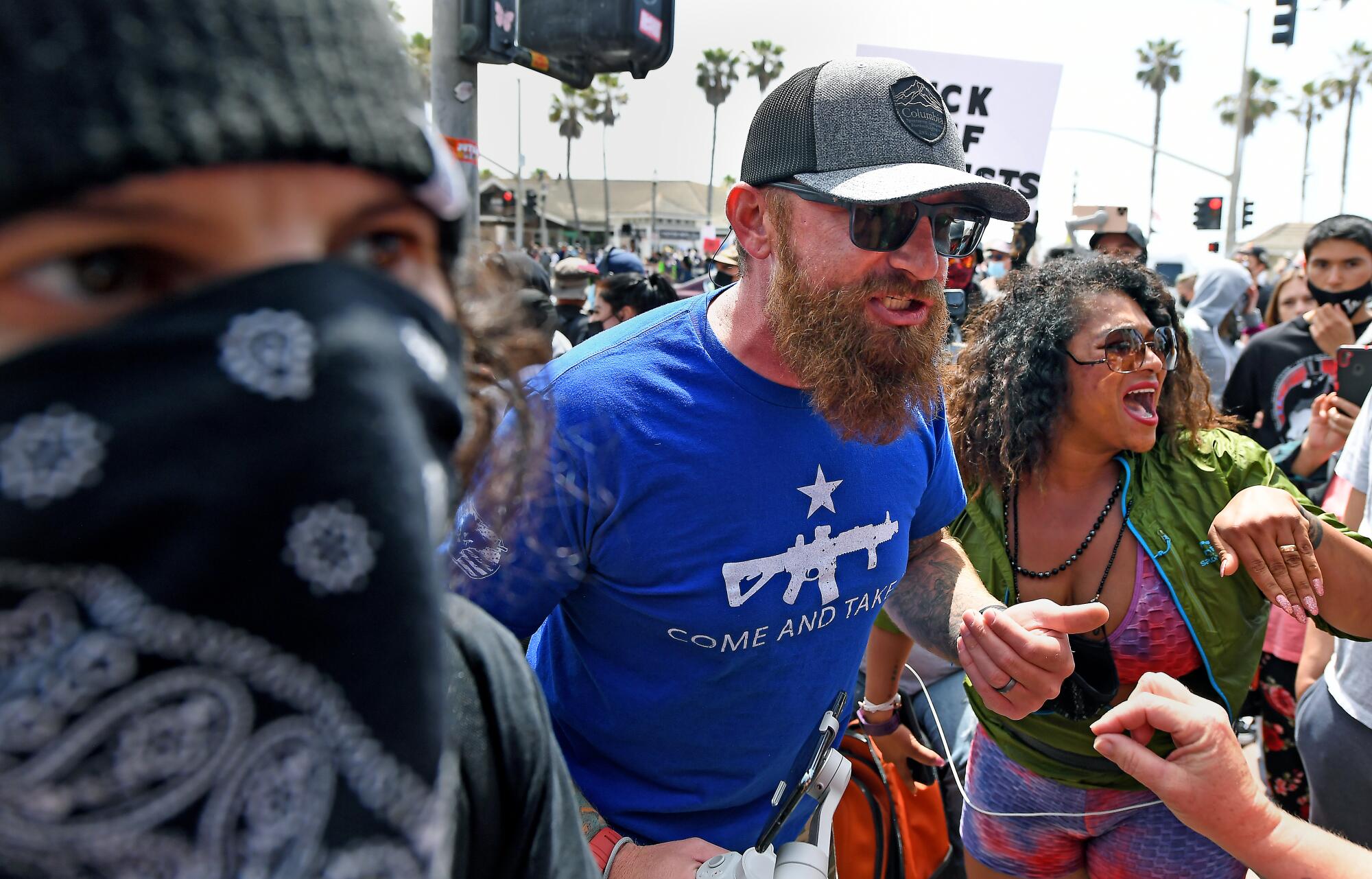 A bearded man with a hat, sunglasses and a T-shirt with a gun graphic on it argues amid a crowd.
