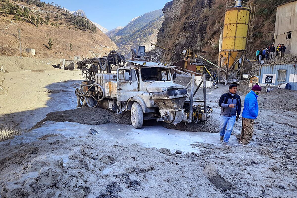 Two men stand near a damaged truck amid hilly terrain.
