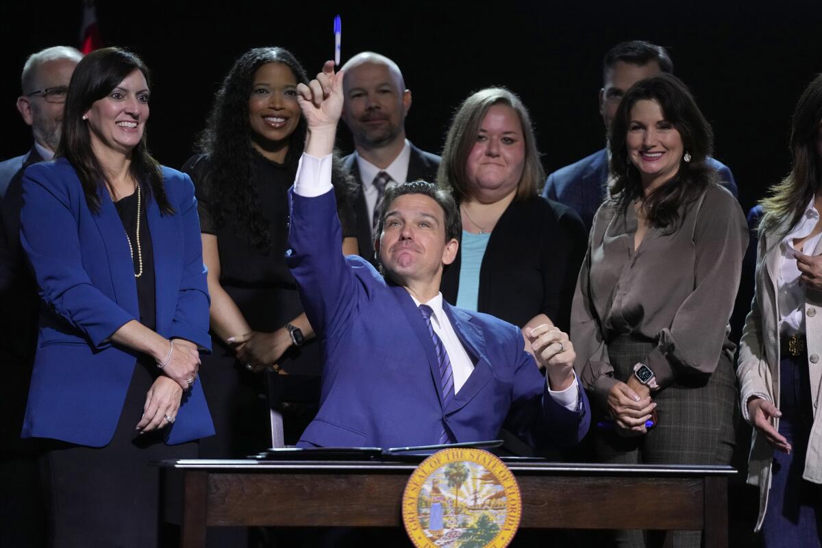 Florida Gov. Ron DeSantis, sitting in front of several supporters at a desk with the state seal, throws a pen to someone