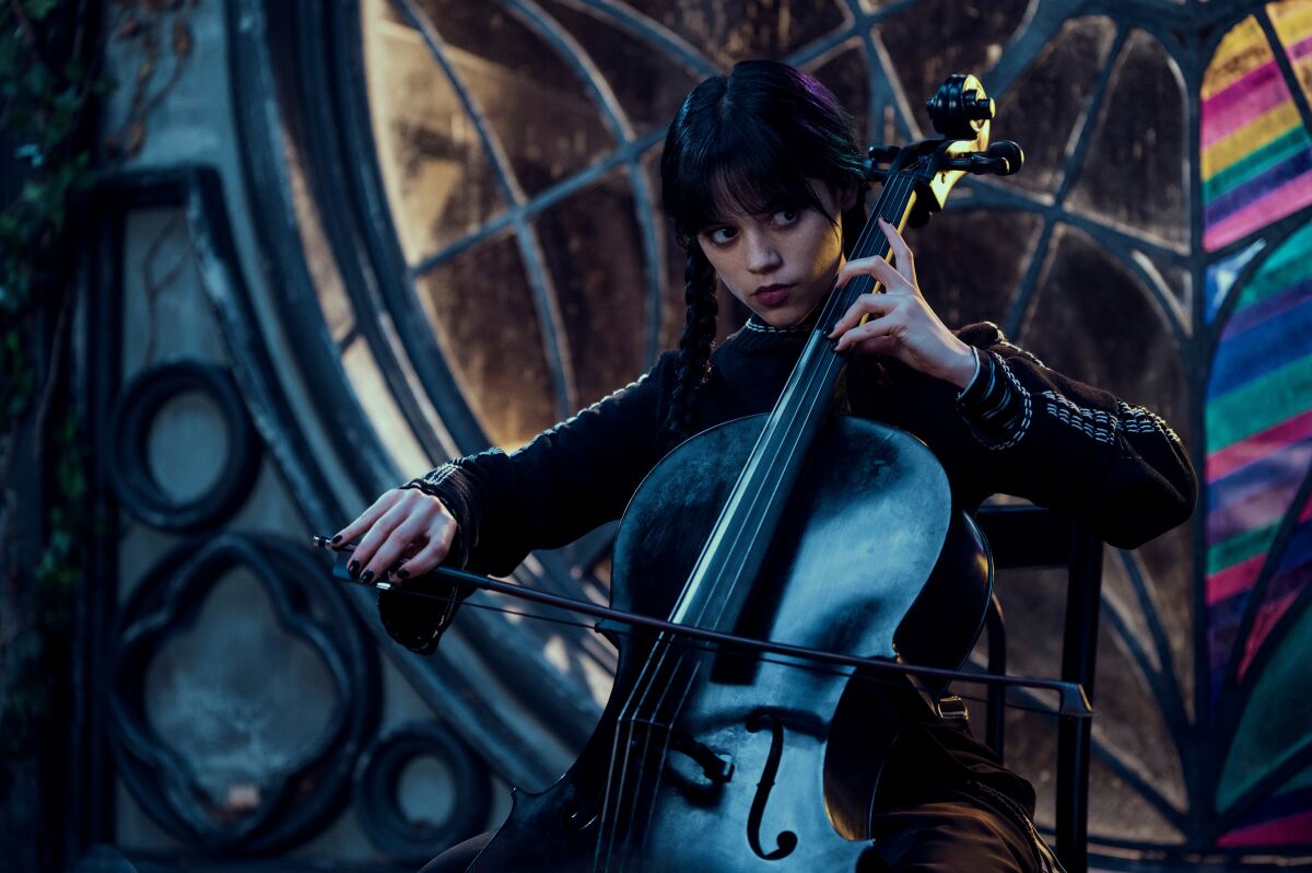 A young woman dressed in black and wearing pigtails plays the cello in front of a window