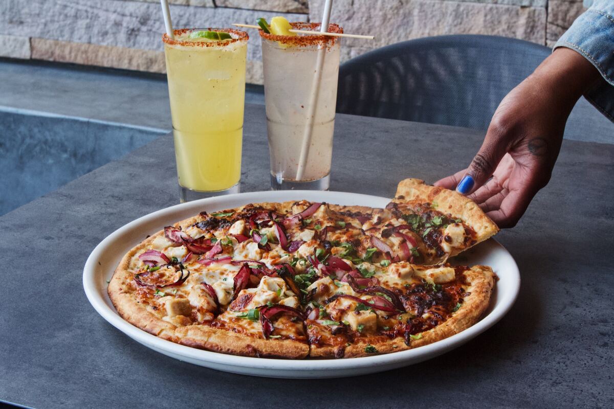 Two cocktails, one clear and one yellow, behind a barbecued chicken pizza. From the right, a hand pulls a slice out.