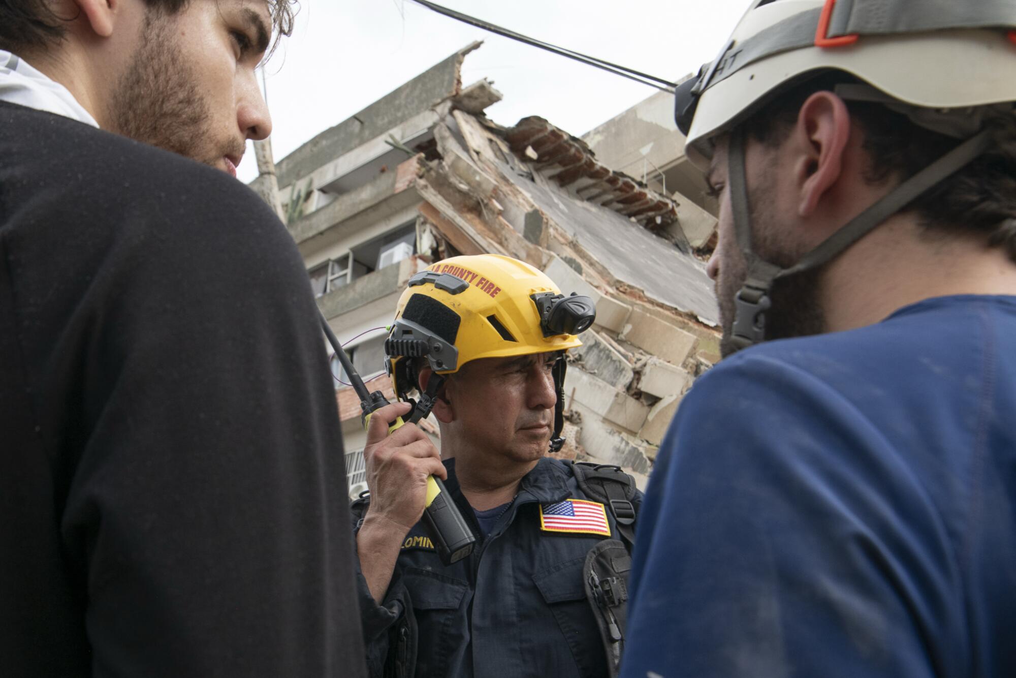 A man in a yellow hard hat and dark clothes with a patch of the U.S. flag, center, holds a walkie-talkie