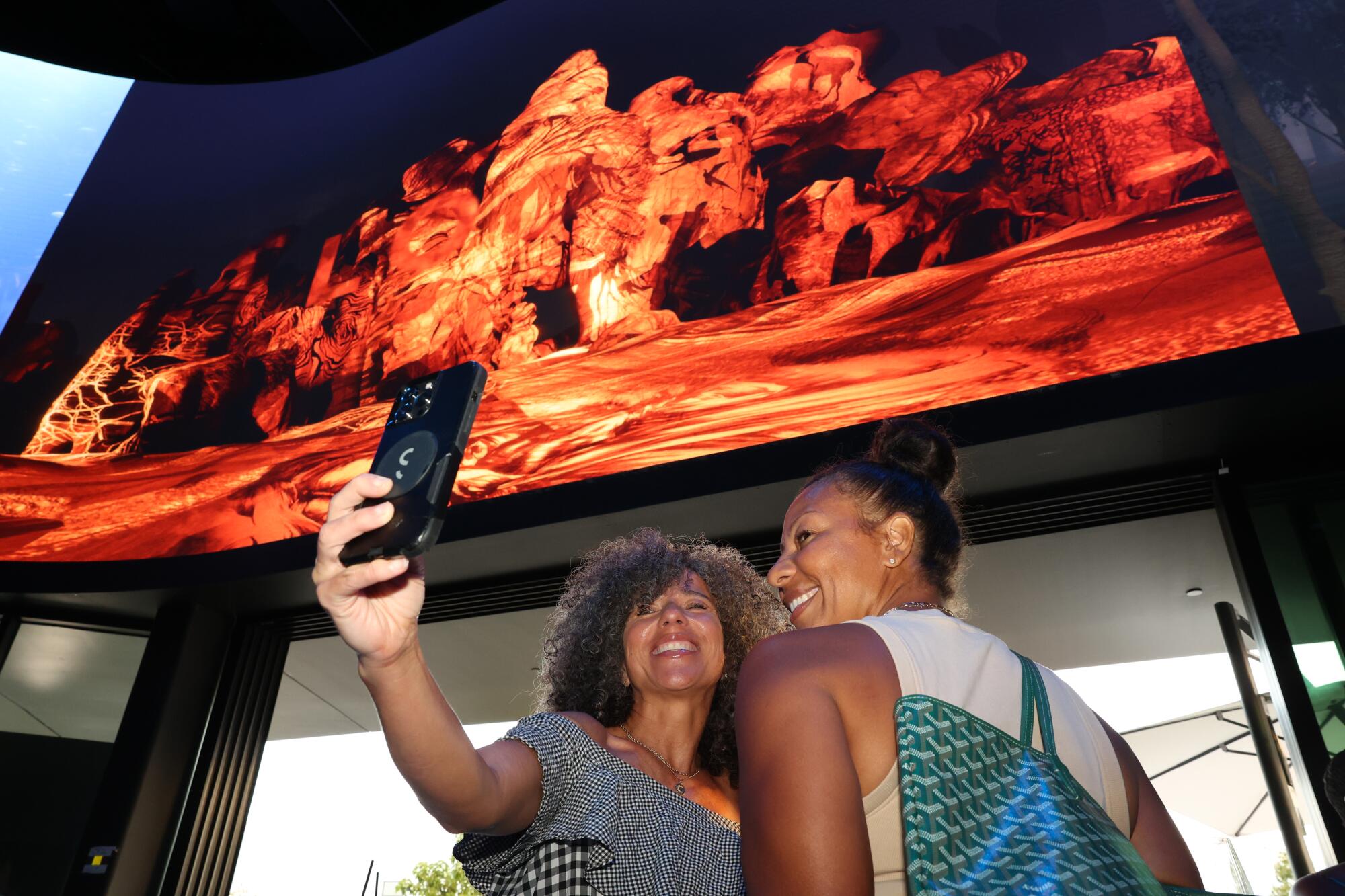 Two women take a selfie below a large screen.