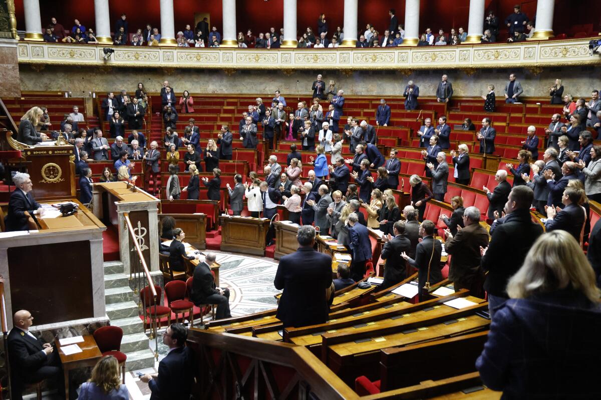 Members of Parliament stand and applaud in a semicircular auditorium with red seats.