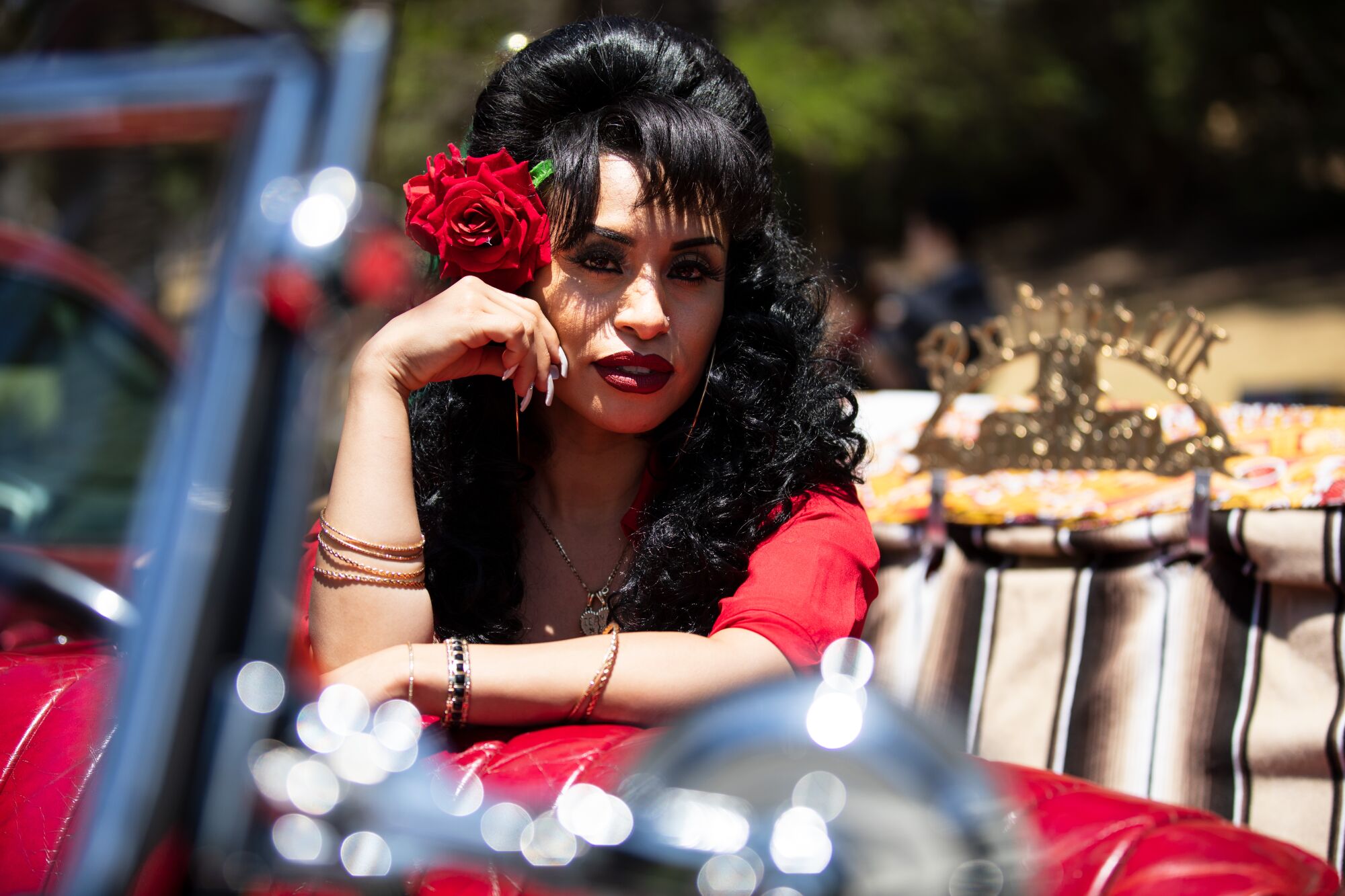 A woman with a bright red flower in her dark hair leans an elbow on a car's red leather seat.