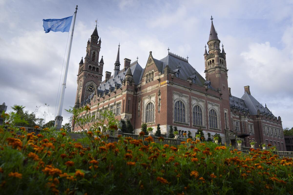 A view of a grand brick building with two towers and a gray roof and a blue flag flying nearby and flowers in the foreground