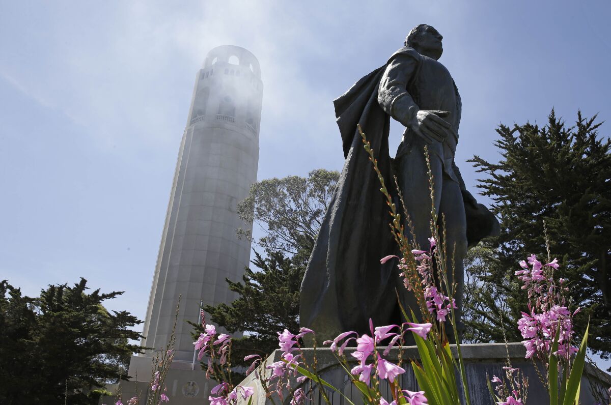 A statue stands in the shadow of a cylindrical-shaped tower, with pink flowers in the foreground