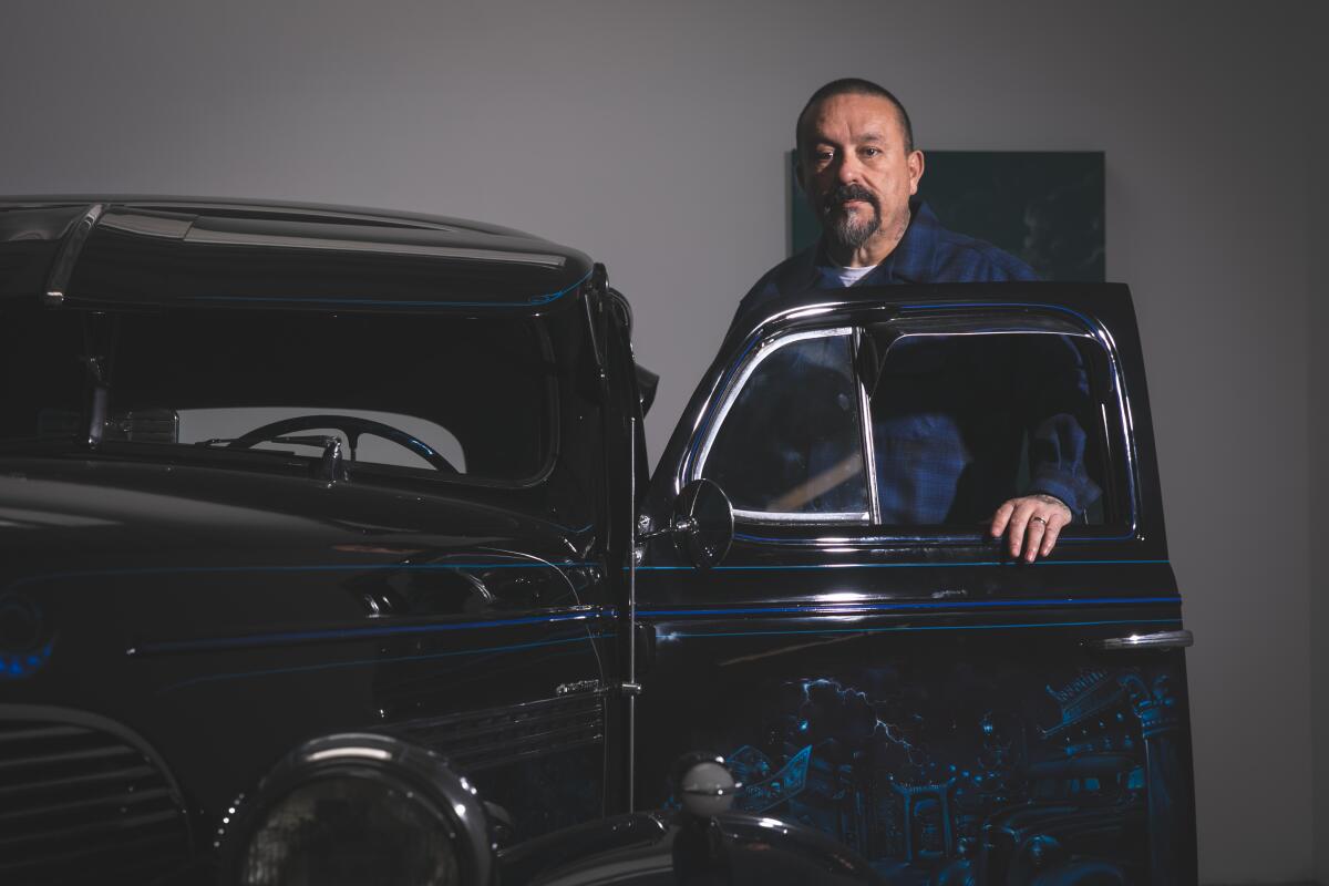 Mister Cartoon, wearing a blue Pendleton and a goatee, stands next to a custom car that has the driver side door open.