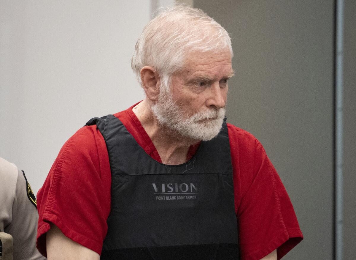 A bearded man wearing a red shirt and a bullet-proof vest enters court.