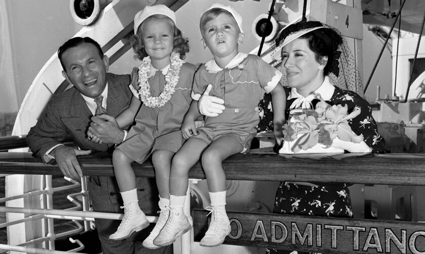 A smiling man and woman stand at a ship's rail, where two small children in matching outfits are perched.