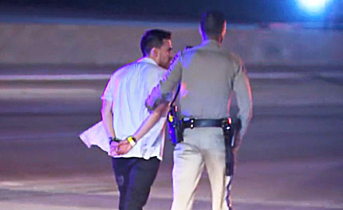 A law enforcement officer guides a handcuffed man on the side of a freeway